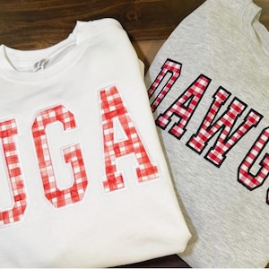 May include: Two sweatshirts, one white and one gray, are displayed. The white sweatshirt has "UGA" in red and white gingham letters. The gray sweatshirt has "DAWGS" in the same gingham pattern. Both sweatshirts are folded.