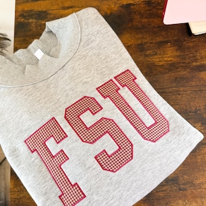May include: A light gray crewneck sweatshirt featuring the letters FSU in a collegiate font. The letters are filled with a red and white gingham pattern and are outlined with a thick red stitched border. The fabric has a soft, heathered texture.