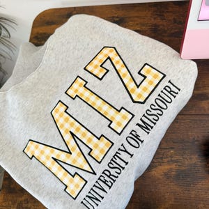 May include: A light gray crewneck sweatshirt featuring the letters MIZ in a yellow and white gingham pattern with a black outline, accompanied by the text UNIVERSITY OF MISSOURI printed in black lettering underneath the large letters.