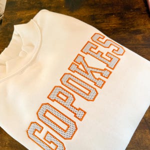 May include: A cream-colored crewneck sweatshirt featuring the word GOPOKES embroidered in large, block-style letters. The lettering has a bright orange outline and a white interior filled with a pattern of small black dots.