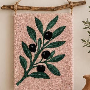 May include: A rectangular wall hanging featuring a textured light pink tufted background with a central embroidered design of a green olive branch with dark black olives. The piece is suspended from a natural wooden branch with jute twine.