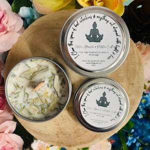 May include: Two silver candle tins with white lids. The candle tins are labelled "I Clear This Space of Bad Intentions & Anything Not Aligned With Light & Love." The text also includes "Intuition, Affirmation & Meditation Candle" and "Clary Sage, Rosemary, Eucalyptus, White Sage". One tin is open and shows a white candle with dried herbs and crystals.