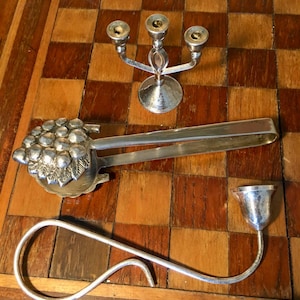 Op de afbeelding: Een set van drie zilveren serveerstukken, waaronder een tang met een druivenmotief, een kleine kan met een tuit en een kandelaar met drie armen.