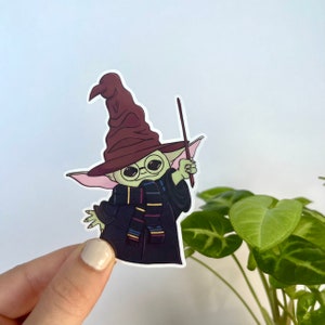 Op de afbeelding: Een sticker met een groen, buitenaards personage met een bordeauxrode tovenaars hoed, een ronde bril en een gestreepte sjaal. Het personage houdt een toverstaf vast en draagt een zwarte toga. De sticker heeft een witte rand.