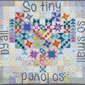 May include: A square patchwork quilt featuring a central heart design composed of various colourful fabric squares and star patterns. The quilt is bordered by the embroidered grey text: So tiny, so small, so loved, by all, in a continuous script.