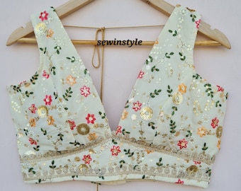 Puede incluir: Una blusa blanca sin mangas con un escote en V profundo. La blusa está decorada con bordados florales y lentejuelas. El escote está adornado con un encaje dorado.