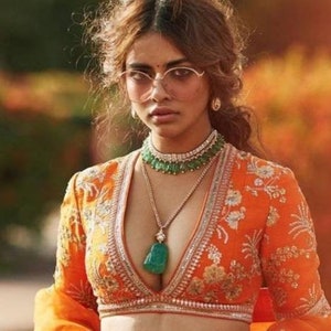 May include: A woman wearing a bright orange top with intricate embroidery and a deep V-neckline. She is wearing a layered necklace with a large green gemstone pendant and a green and diamond choker.