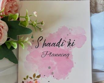 May include: A white planner with the text Shaadi ki Planning printed in black script. The cover features a pink watercolour splash design, small green floral accents, and a cluster of pink roses with green leaves at the bottom left corner.