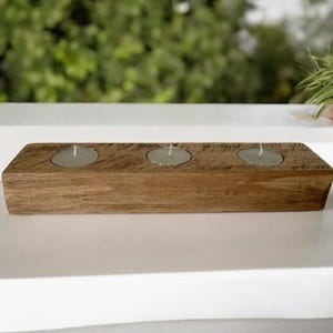 May include: A wooden candle holder with three circular openings for tealight candles. The holder is made of rustic brown wood with a natural finish.