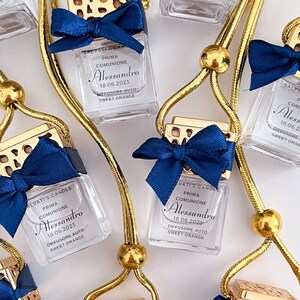 Op de afbeelding: Een set van zes gouden en blauwe autoluchtverfrissers met de tekst "SALVATI'S CANDLE" en "PRIMA COMUNIONE Alessandro 18.06.2023 DIFFUSORE AUTO SWEET ORANGE" erop gedrukt. De luchtverfrissers hangen aan gouden koorden met gouden kralen.