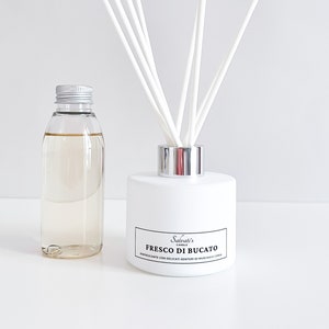 Puede incluir: Botella difusora blanca con tapa plateada y una etiqueta que dice "Salvati's Candles Fresco Di Bucato Diffusore Elegante con Delicati Sentori di Muschio e Limone". La botella está llena de un líquido transparente y tiene varillas difusoras blancas.