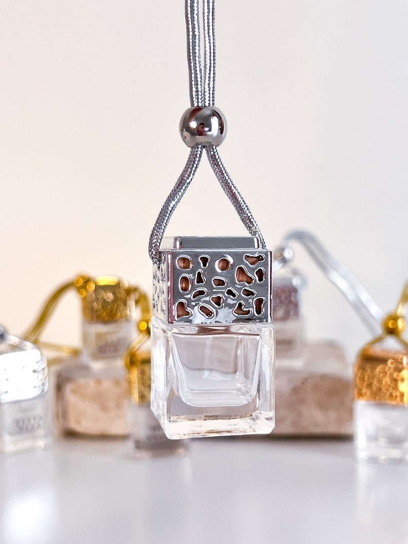 May include: A silver car air freshener with a decorative metal cage and a clear glass bottle. The air freshener is hanging from a silver cord with a silver bead.