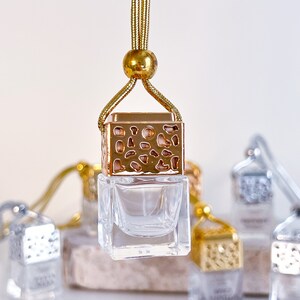 May include: Gold car air freshener with a clear glass bottle and a gold metal cage design. The air freshener is hanging from a gold cord.