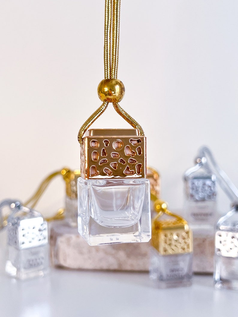 May include: Gold car air freshener with a clear glass bottle and a gold metal cage design. The air freshener is hanging from a gold cord.