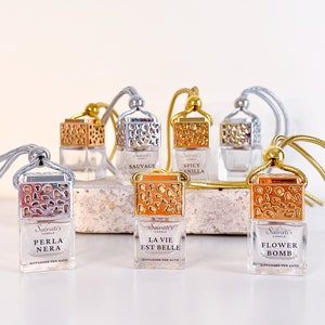 May include: Six glass car air freshener bottles with gold and silver caps. The bottles are filled with a clear liquid and have a variety of scents, including "Perla Nera", "La Vie Est Belle", "Flower Bomb", "Sauvage", and "Spicy Vanilla".