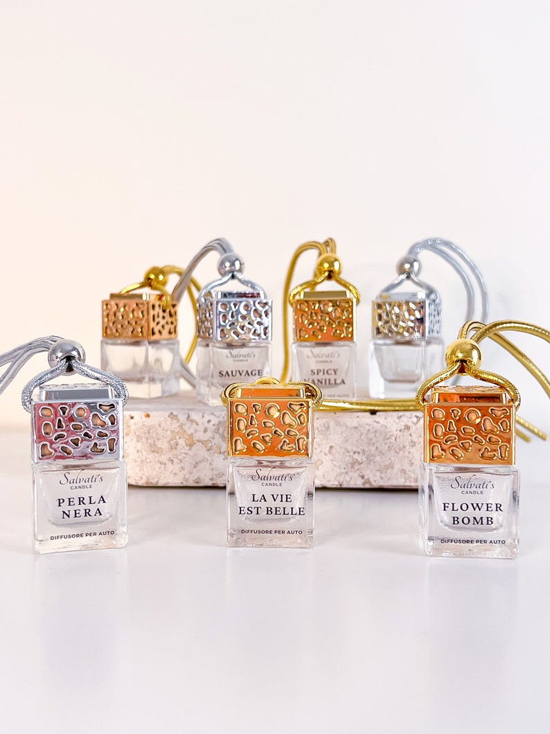 May include: Six glass car air freshener bottles with gold and silver caps. The bottles are filled with a clear liquid and have a variety of scents, including "Perla Nera", "La Vie Est Belle", "Flower Bomb", "Sauvage", and "Spicy Vanilla".