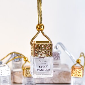 May include: Gold car air freshener with a gold chain and a clear glass bottle. The bottle has a gold lid and a label that says "Salvati's Candle Spicy Vanilla Diffusore Per Auto".