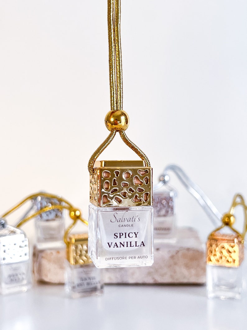 May include: Gold car air freshener with a gold chain and a clear glass bottle. The bottle has a gold lid and a label that says "Salvati's Candle Spicy Vanilla Diffusore Per Auto".