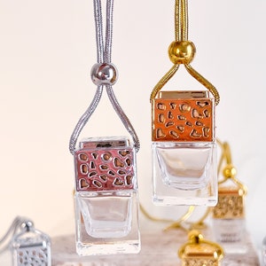 May include: Two car air fresheners with gold and silver chains. The air fresheners are square glass bottles with a decorative metal top. The metal top has a geometric pattern and is in a rose gold color. The bottles are empty and ready to be filled with essential oils.