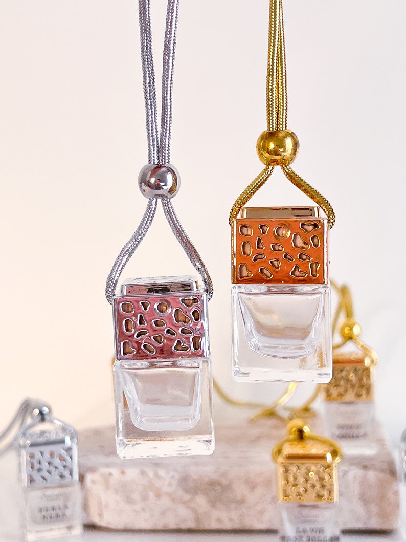 May include: Two car air fresheners with gold and silver chains. The air fresheners are square glass bottles with a decorative metal top. The metal top has a geometric pattern and is in a rose gold color. The bottles are empty and ready to be filled with essential oils.
