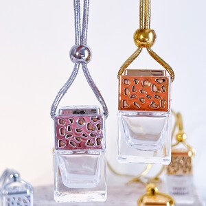 May include: Two car air fresheners with clear glass bottles and decorative metal tops. The tops are silver and gold with a geometric pattern. The fresheners are hanging from silver and gold cords.
