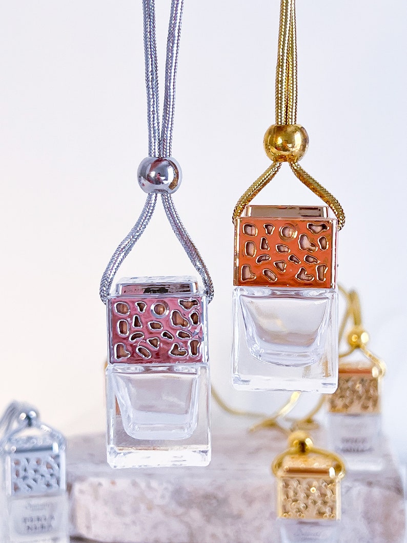 May include: Two car air fresheners with clear glass bottles and decorative metal tops. The tops are silver and gold with a geometric pattern. The fresheners are hanging from silver and gold cords.