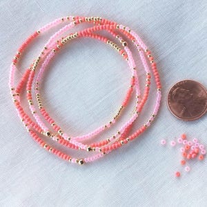May include: A coiled beaded bracelet featuring alternating light pink, coral, and metallic gold beads. A copper penny is placed next to the bracelet for scale, along with a small pile of loose pink and coral beads scattered on the surface.