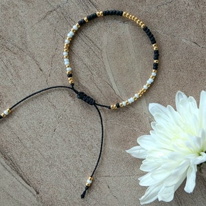 May include: A black and white beaded bracelet with gold accents. The bracelet is tied with a black cord and has a sliding knot for adjusting the size.