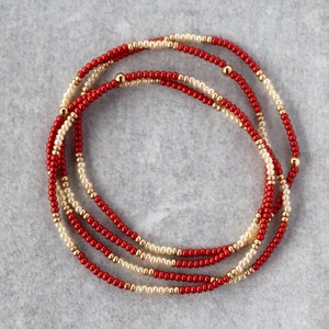 May include: A red and cream beaded necklace with gold accents. The necklace is made of small beads and is layered in a circular pattern.