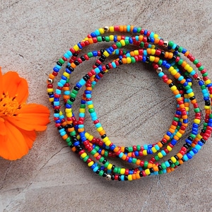 May include: A collection of colorful beaded waist beads in a circular arrangement. The beads are in various colors, including red, yellow, blue, green, and black. An orange flower with a green stem is next to the beads.
