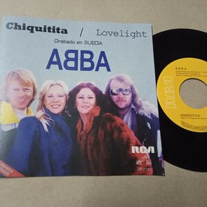 Puede incluir: Un disco de vinilo de 45 RPM de ABBA "Chiquitita / Lovelight". La funda del disco presenta una foto de los miembros de la banda y el logotipo de RCA Victor. El disco es negro con una etiqueta amarilla.