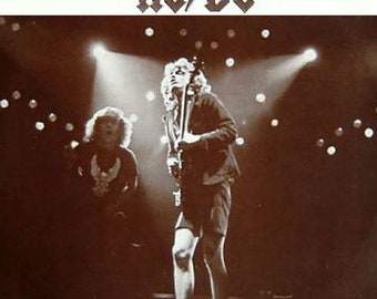 May include: A black and white photo of a rock band performing on stage. The band is AC/DC, and the photo shows two band members, one playing a guitar and the other standing next to him. The photo is taken from a low angle, and the stage lights are visible in the background.
