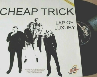 May include: A black and white promotional record album cover for the band Cheap Trick. The album is titled "Lap of Luxury" and features a photo of the band members in black and white. The album cover also includes the Epic Records logo and the text "Lo que se ve y se escucha en el Mexico moderno Disco promocional".