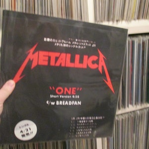 May include: A black vinyl record with a red Metallica logo and the text "ONE" Short Version 4:38 c/w BREADFAN. The record is in a plastic sleeve and is on a shelf with other records.