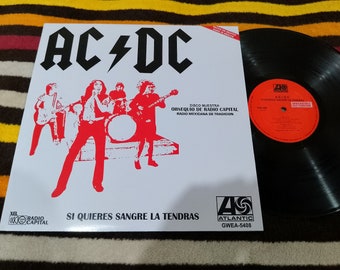 May include: A vinyl record and album cover featuring the band AC/DC. The album cover is white with red graphics of the band members and the band's logo. The text "SI QUIERES SANGRE LA TENDRAS" is visible. The record is black.