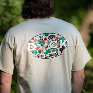 May include: A beige t-shirt with a camouflage oval graphic on the back. The graphic features a duck in flight and the text "BIG DUCK ENERGY".