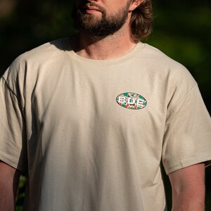 May include: A beige t-shirt with a green, white and orange oval patch that says "BDE" on it.