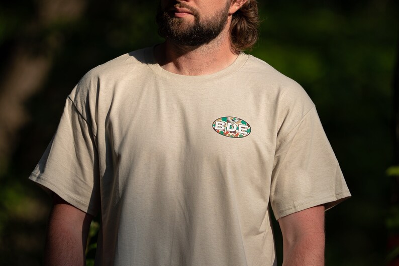 May include: A beige t-shirt with a green, white and orange oval patch that says "BDE" on it.