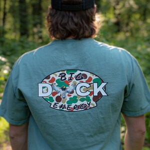 May include: A teal blue t-shirt with a white oval graphic on the back. The graphic features a camouflage pattern with a duck flying in the centre. The text "BIG DUCK ENERGY" is printed in white around the graphic.