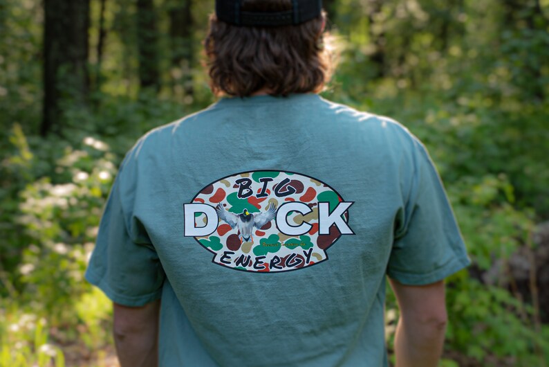 May include: A teal blue t-shirt with a white oval graphic on the back. The graphic features a camouflage pattern with a duck flying in the centre. The text "BIG DUCK ENERGY" is printed in white around the graphic.