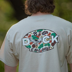May include: A beige t-shirt with a camouflage oval graphic that says "BIG DUCK ENERGY" in white letters.