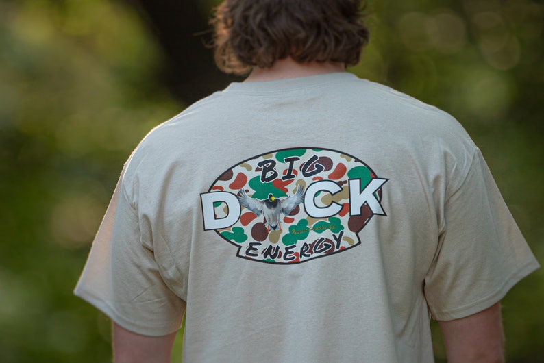 May include: A beige t-shirt with a camouflage oval graphic that says "BIG DUCK ENERGY" in white letters.