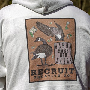 May include: White hooded sweatshirt with a brown rectangular graphic featuring two geese and the text "Bands Make Her Dance" and "Recruit Creative Co."