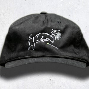 May include: Black baseball cap with a white embroidered design of a deer jumping over an arrow. The cap has a curved brim and an adjustable strap. The design is a simple, line-art style.