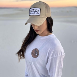 May include: A tan mesh trucker hat with a white patch that says "RECRUITS" with a black antler design. The hat is worn by a person wearing a white long-sleeve shirt with a brown patch on the front that says "RECRUITS" with a black antler design.