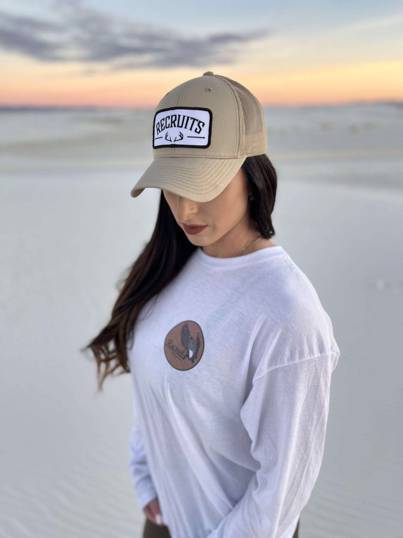 May include: A tan mesh trucker hat with a white patch that says "RECRUITS" with a black antler design. The hat is worn by a person wearing a white long-sleeve shirt with a brown patch on the front that says "RECRUITS" with a black antler design.