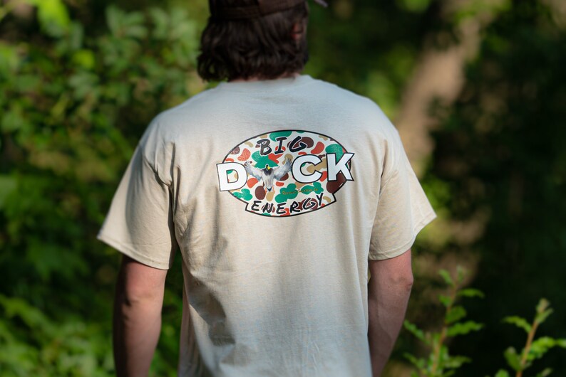 May include: A beige t-shirt with a camouflage oval graphic on the back. The graphic features a duck in flight and the text "BIG DUCK ENERGY".