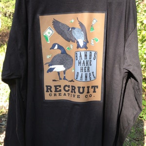 May include: Black long-sleeve t-shirt with a brown rectangle graphic featuring two geese flying with money falling around them. The graphic also includes the text "Bands Make Her Dance" and "Recruit Creative Co."
