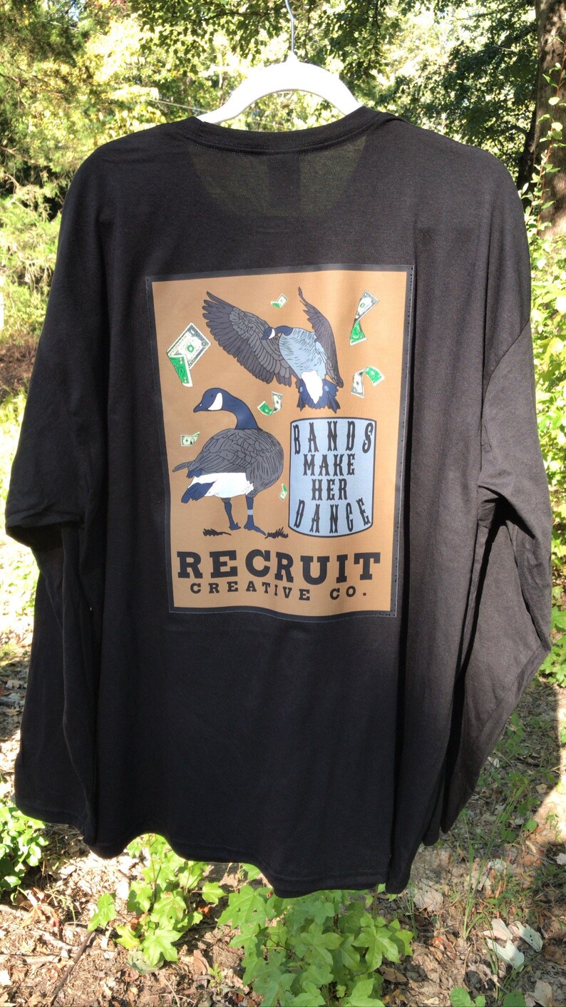 May include: Black long-sleeve t-shirt with a brown rectangle graphic featuring two geese flying with money falling around them. The graphic also includes the text "Bands Make Her Dance" and "Recruit Creative Co."