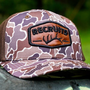May include: A brown and white camouflage trucker hat with a black leather patch that says "RECRUITS" with a brown antler design.